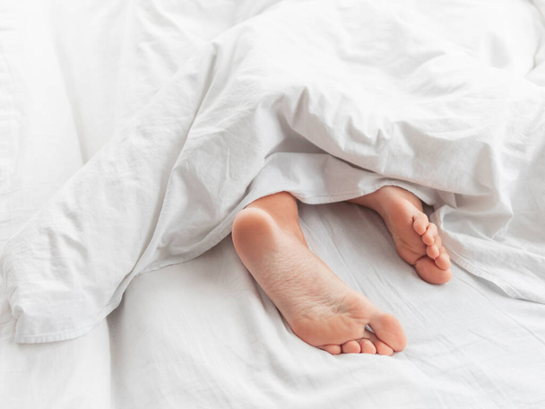 View on bare feet under blanket. Bedroom lit with morning light. Woman sleeps with comfort. White crumpled linen. - Photo, image