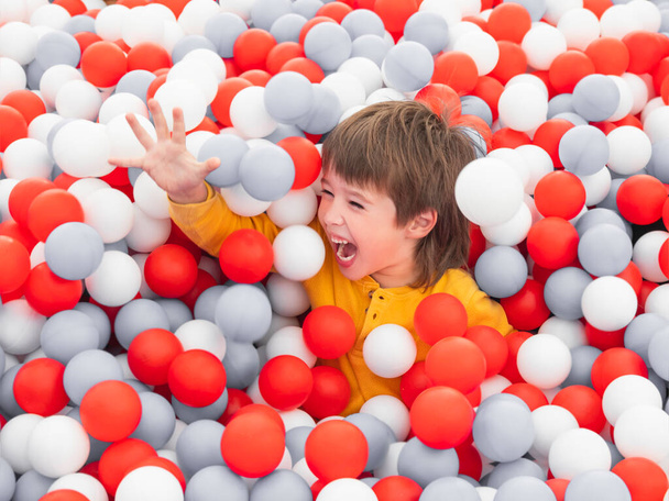 Laughing boy plays in ball pit. White and red plastic balls in dry paddling pool. Leisure activity in playroom at kindergarten. Leisure activity. Active recreation. - Photo, Image