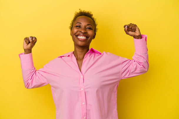 Young african american woman isolated on yellow background showing strength gesture with arms, symbol of feminine power - Photo, Image