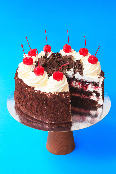 Sliced Birthday cake on the wooden cake stand. Beautiful chocolate sponge cake decorated with Maraschino Cherries (cocktail cherry) and whipped cream. Blue background. Copy space. Food photography for recipe. - Photo, Image