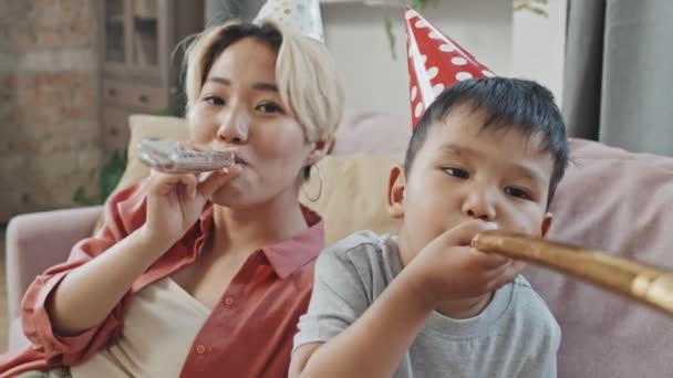 Medium close-up met slowmo van vrolijke Aziatische moeder en zoontje dragen grappige verjaardag hoeden blazen partij hoorns op camera vieren verjaardag thuis - Video