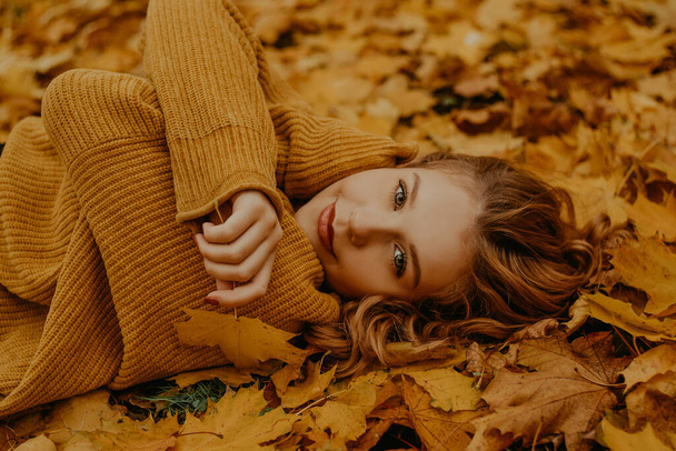 Krásná dívka leží v parku na podzim. Puberťák ve žlutém svetru na pozadí pomerančových listů. Blondýnka se dívá do kamery. Móda a styl. - Fotografie, Obrázek