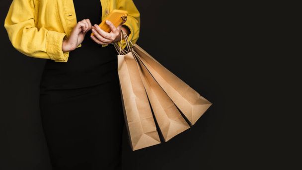 Uma mulher usa seu telefone e segura sacos de compras Black Friday enquanto está de pé contra um fundo preto. - Foto, Imagem