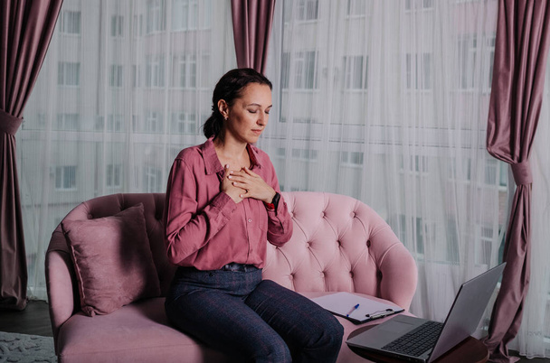 une femme en chemise rose s'assoit sur le canapé et mène une consultation à distance à l'aide d'un ordinateur portable - Photo, image