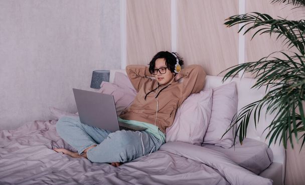 девочка-подросток сидит на кровати и удаленно общается через ноутбук и наушники - Фото, изображение