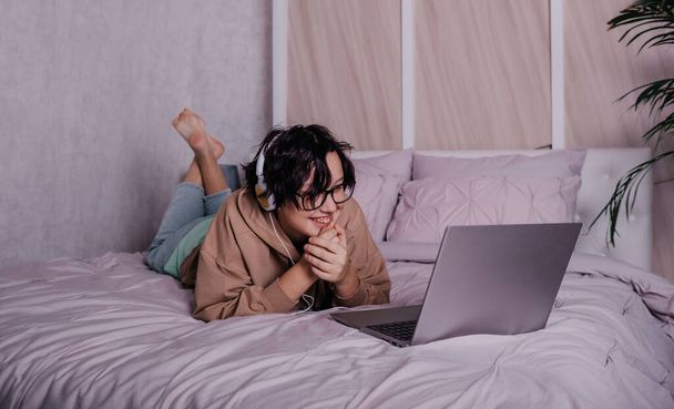 egy nevető tini lány fülhallgatóval és egy laptop fekszik az ágyon a szobában. - Fotó, kép