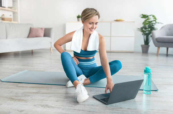 Femme souriante assise sur un tapis de yoga à l'aide d'un ordinateur portable - Photo, image