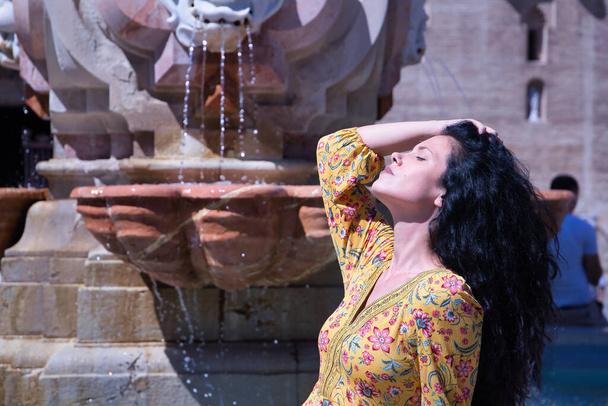 Middle-aged adult Hispanic woman with black curly hair, wearing a colorful outfit, sitting on a fountain, touching her hair on a sunny day. Concept hair, hairdresser, beauty, shampoo, sun. - Photo, Image