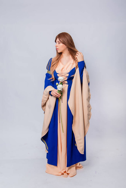 An attractive woman with long hair in a medieval, fantasy, blue and beige dress with long, large sleeves, posing with white roses in her hands, isolated on a white background. - Foto, imagen