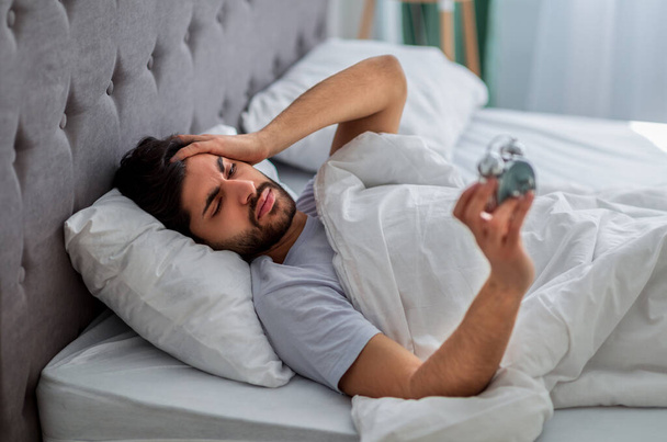 Unhappy arab man lying in bed with alarm clock, not willing to get up in morning and suffering from headache - Photo, Image