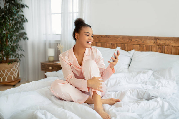 Feliz señora afroamericana utilizando el teléfono inteligente navegar por Internet y mensajes de texto en el teléfono sentado en la cama en casa - Foto, imagen
