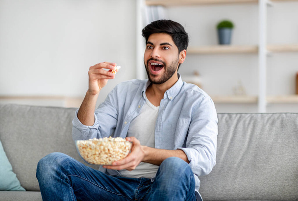 Retrato del hombre árabe emocional viendo la televisión y comiendo palomitas de maíz, descansando en el sofá en casa, espacio libre - Foto, imagen