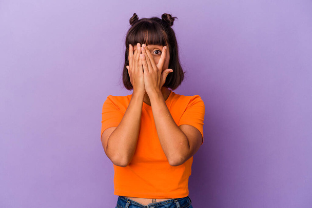 Jeune femme métisse isolée sur fond violet cligner des yeux à travers les doigts effrayé et nerveux. - Photo, image