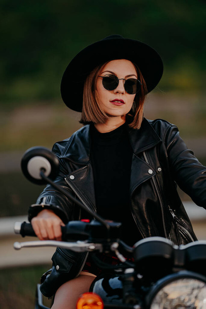 Close-up of portrait of stylish woman in hat on classic black retro-styled motorcycle while standing on road. Sexy lady in leather jacket. Bike, driver culture concept. - Foto, imagen