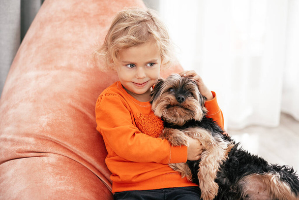 portrait of pretty little girl sitting on orange chair with dog in arms looking at side and smiling - Foto, Imagem