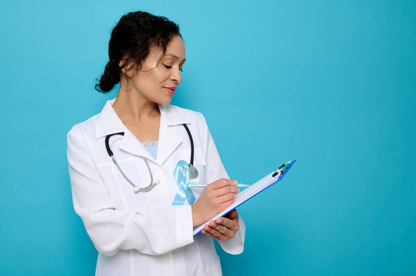 Middle aged mixed race beautiful female doctor wearing a blue satin ribbon, symbolic bow color raising awareness in diabetes day , makingtes on a clipboard, isolated over blue background, copy space - Photo, Image