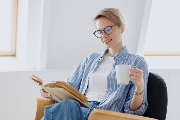 a young European woman with short blonde hair is drinking coffee or tea and reading a book - Zdjęcie, obraz