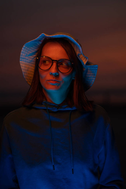 a European woman illuminated by neon light in a hat against the background of the sunset sky. neon lights and a woman at sunset. stylish cyber photo. color filters on neon lamps. creative photo and - Zdjęcie, obraz