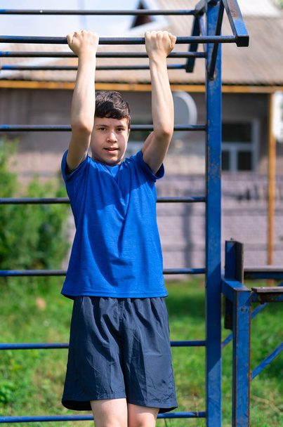 teenage boy exercising outdoors, sports ground in the yard, he pulls himself up on the horizontal bar, healthy lifestyle - Photo, Image