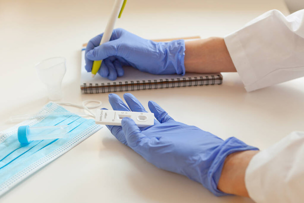 sars covid antigen test. maska a laboratorní sklo u stolu. lékař ruce v modrých rukavicích drží rychlý test a psaní výsledků na vědomí - Fotografie, Obrázek
