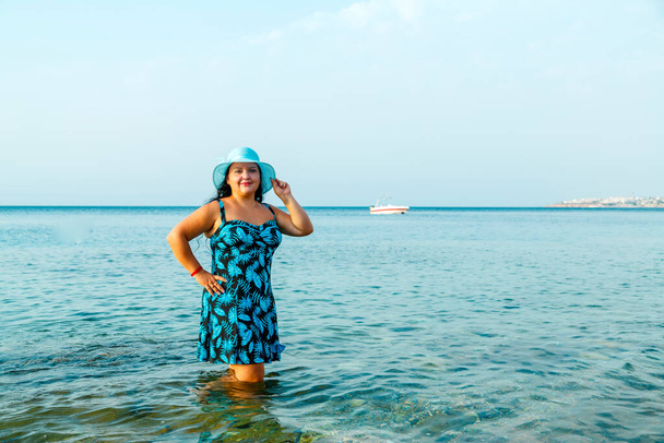 A happy Jewish woman in a blue sundress and hat stands in the sea knee-deep in water. Horizontal photo - Foto, Imagen