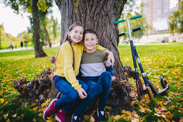 Друзья дети счастливо позируют, сидя под деревом в осеннем парке рядом со скутерами. Счастливые дети. Эко-транспорт. Близнецы на природе. Брат и сестра смеются в объятиях природы. - Фото, изображение