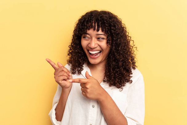 Young mixed race woman isolated on yellow background points with thumb finger away, laughing and carefree. - Photo, Image