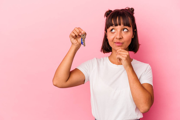Молодая женщина смешанной расы держит ключи изолированы на розовом фоне глядя в сторону с сомнительным и скептическим выражением. - Фото, изображение