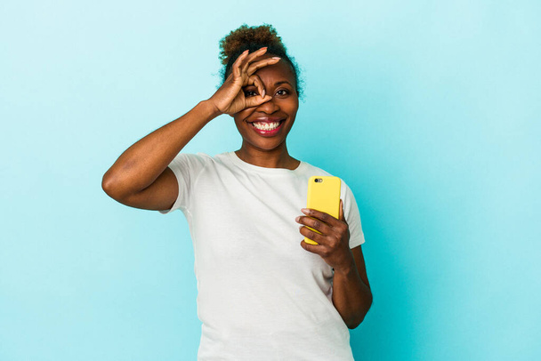 Jovem afro-americana segurando um telefone celular isolado no fundo azul animado mantendo ok gesto no olho. - Foto, Imagem