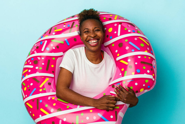 jong Afrikaans amerikaans vrouw met opblaasbare donut geïsoleerd op blauwe achtergrond lachen en plezier hebben. - Foto, afbeelding