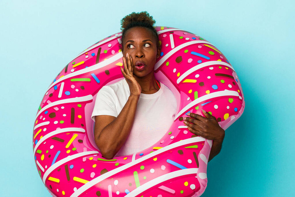 Jonge Afro-Amerikaanse vrouw met opblaasbare donut geïsoleerd op blauwe achtergrond zegt een geheim warm remnieuws en kijkt opzij - Foto, afbeelding