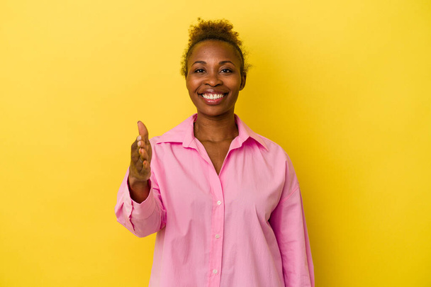 Young african american woman isolated on yellow background stretching hand at camera in greeting gesture. - Photo, Image