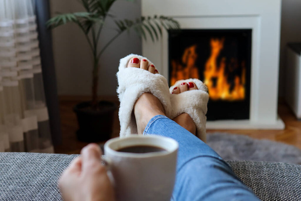 Γυναικεία γυμνά πόδια σε λευκό δωμάτιο παντόφλες και ζεστά πόδια δίπλα στο τζάκι. Μια γυναίκα ξαπλώνει στο κρεβάτι και χαλαρώνει με ένα ζεστό ρόφημα καφέ - Φωτογραφία, εικόνα