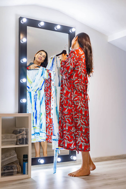 Young brunette woman choosing dress in front on mirror at dressing room getting ready to party event - 写真・画像