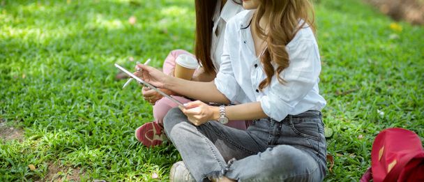 Zugeschnittenes Bild zweier enger Freundinnen, die sich am Wochenende gemeinsam in einem Park entspannen. Einsatz eines Tablet-Geräts - Foto, Bild