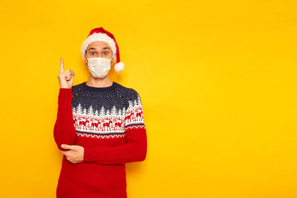 sokkolt, izgatott férfi karácsonyi pulóverben rénszarvas, orvosi maszk, újévi Mikulás kalap, mutatja mutatóujját felfelé, jó ötlet jött. izolált sárga háttér szöveges hellyel. nyaralás - Fotó, kép