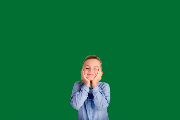 Счастливый малыш держит руки на плечах, глаза закрыты, улыбается довольным, весело проводит время с друзьями, носит повседневную одежду, изолированную на зеленом фоне.. - Фото, изображение