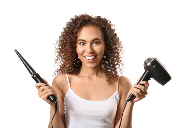 Beyaz arka planda saç kurutma makinesi ve saç maşası olan genç Afro-Amerikalı kadın. - Fotoğraf, Görsel