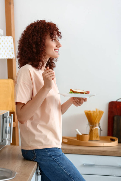 Pretty young woman with tasty sandwich near counter in kitchen - Foto, Imagen