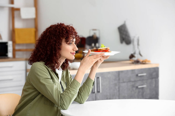 Mutfaktaki masada lezzetli sandviçle tabağı tutan genç ve güzel bir kadın. - Fotoğraf, Görsel