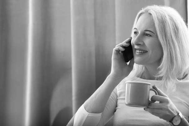 Beautiful mature woman with cup of coffee talking by mobile phone at home - Photo, Image