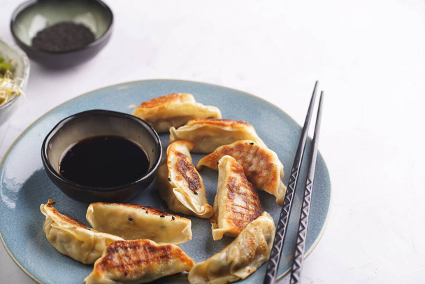Японские пельмени гёдза, фаршированные куриным мясом на голубой тарелке с соевым соусом, микрозелень, семена кунжута и черные палочки для отбивных - Фото, изображение