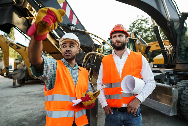 Two men engineers discussing their work standing against construction machines - Foto, Imagen