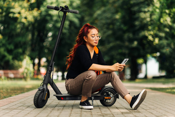 Happy attractive asian female , wearing glasses standing with electric scooter outdoors in park, woman with red backpack using mobile phone - Fotoğraf, Görsel