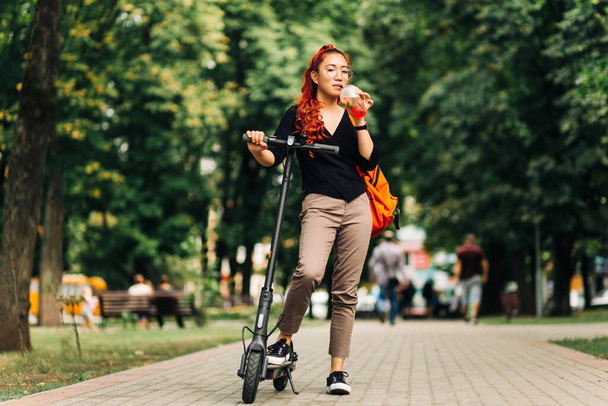 Uśmiechnięta Azjatka, używająca skutera elektrycznego w parku, Nowy gadżet do szybkiego ruchu miejskiego bez korków, Nowoczesna kobieta jeżdżąca na skuterze elektrycznym w parku - Zdjęcie, obraz