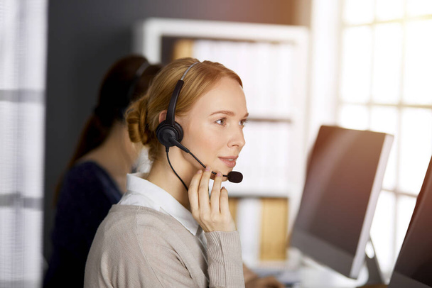 Friendly caucasian businesswoman talking by headset in sunny office. Call center and diverse people group in business - Fotoğraf, Görsel
