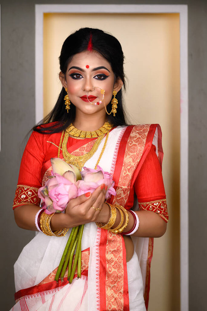 Portrét hezké mladé indiánské dívky v tradičním indiánském saree, zlatých špercích a náramcích, držící v rukou květiny. Indická kultura, náboženství a móda - Fotografie, Obrázek