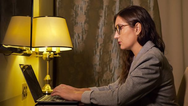 woman in glasses and in a business suit in the evening works at a laptop - Photo, Image
