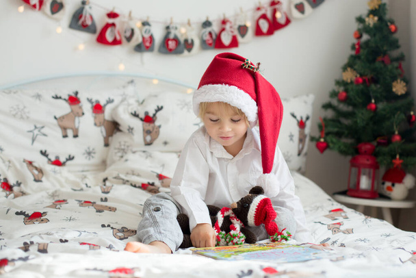 Beautiful toddler child, reading book in bed on Christmas, decoration around him - Photo, image