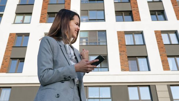 Frau im Businessanzug blickt auf das Telefon und freut sich über den Hintergrund des Gebäudes, - Foto, Bild